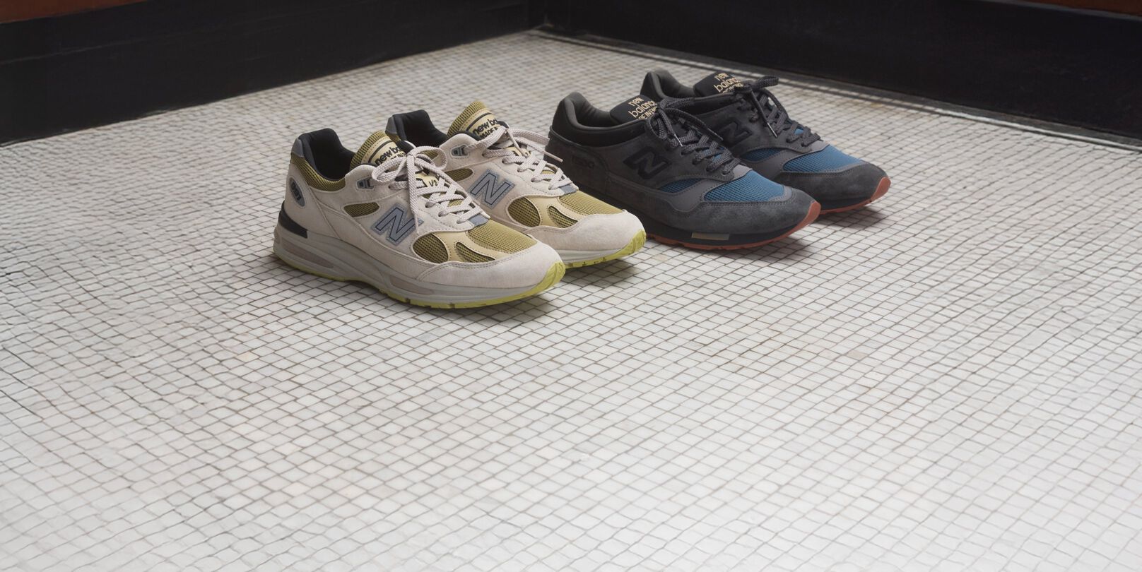 Two pairs of New Balance Made in UK sneakers on a white tiled floor with wooden paneled walls in the background.   