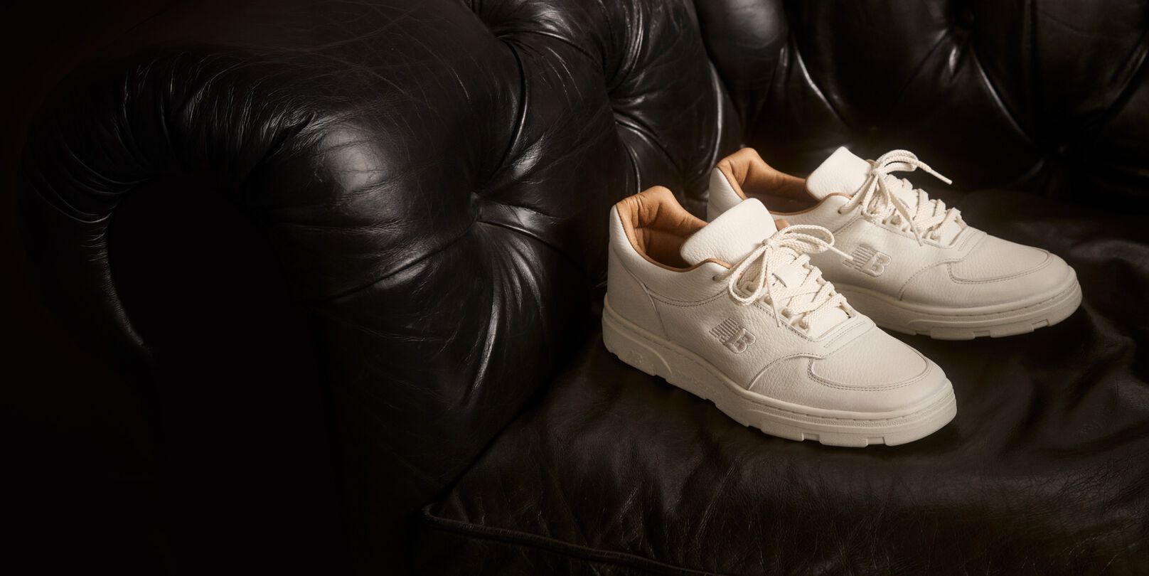 A pair of New Balance Made in UK sneakers sitting on a black leather sofa.