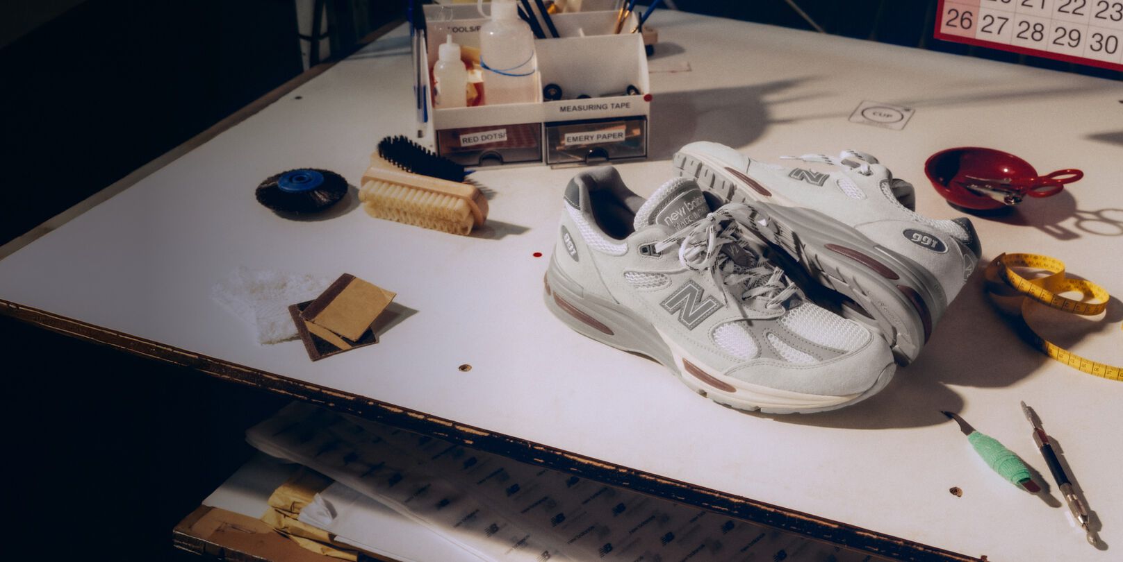 A pair of New Balance Made in UK sneakers on a white workbench surrounded by shoemaking tools and other textile equipment. 