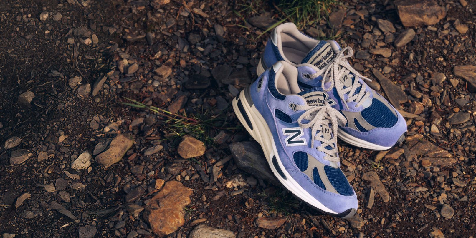 A pair of New Balance Made in UK sneakers on a raw earth surface of dirt and stones. 