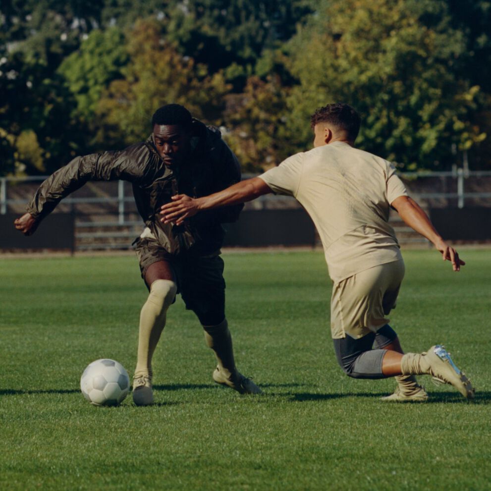 Three people playing soccer on a grassy field with a goal and trees in the background. 