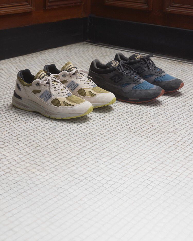 Two pairs of New Balance Made in UK sneakers on a white tiled floor with wooden paneled walls in the background.   